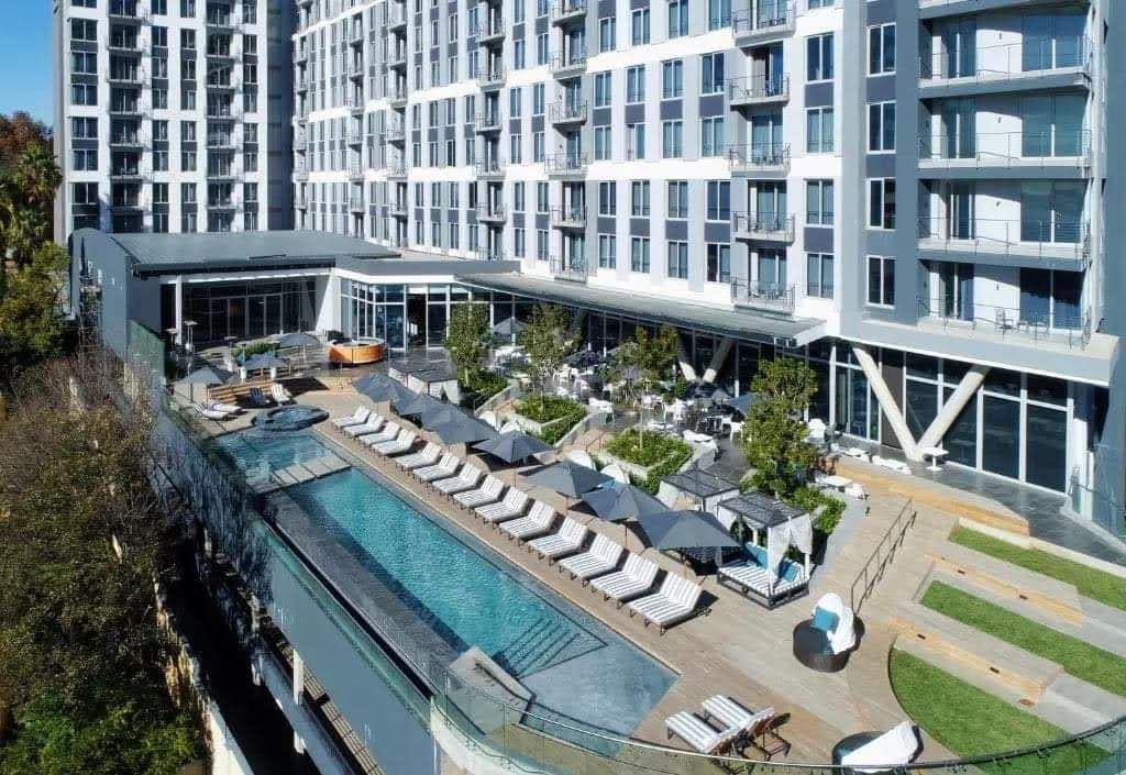 An aerial view of a large outdoor pool area shows numerous sun loungers arranged in rows under shaded umbrellas. Surrounding greenery is visible alongside a spacious patio area, which features seating arrangements. The modern building with large windows stands in the background.