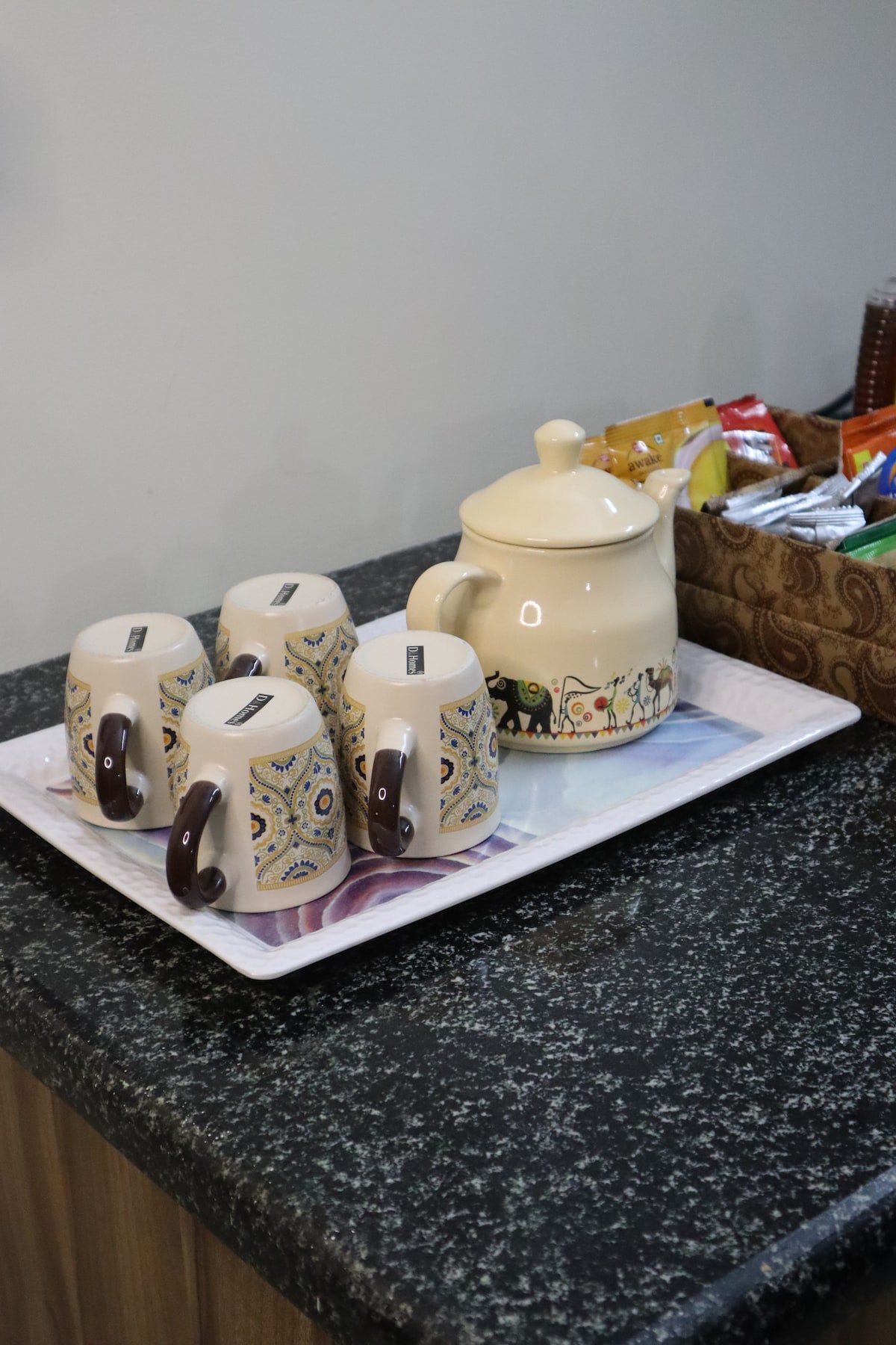 A tray is displayed on a granite countertop, featuring four patterned mugs with handles and a ceramic teapot. A woven basket in the background contains a variety of packaged snacks, contributing to a welcoming refreshment area.