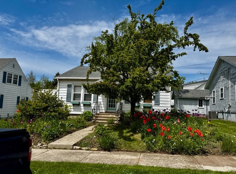 A charming cottage is surrounded by vibrant flower beds featuring tulips and other blooms. A large tree provides shade over the front steps leading to the entrance. The exterior showcases a combination of light and dark siding, with a welcoming front porch.