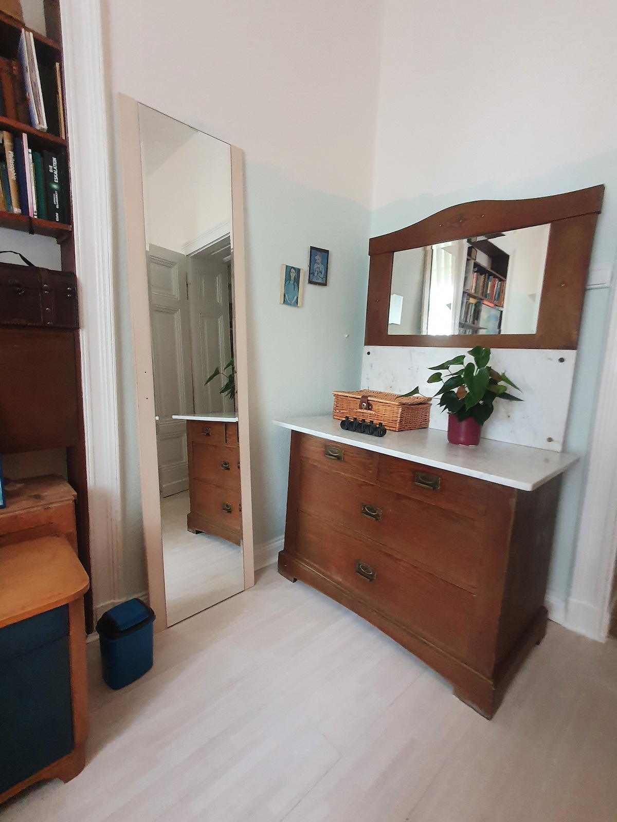 A wooden dresser with a marble top is positioned against a light-colored wall. A mirror is mounted above the dresser, reflecting a small potted plant. To the left, a bookcase filled with books adds a touch of character, while natural light brightens the space.