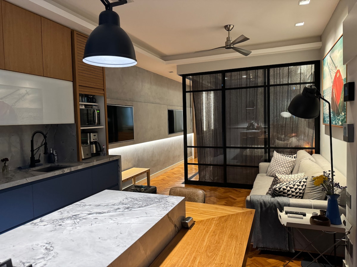 An open-plan living area showcases a modern kitchen with sleek countertops and built-in appliances. A cozy seating area features a light-hued sofa adorned with various textured pillows, complemented by a floor lamp. Large glass panels separate the spaces, enhancing the contemporary design.