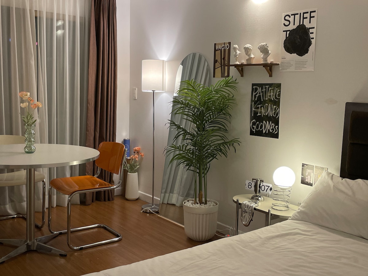 A well-lit room features a spacious bed, a round dining table with two chairs, and a decorative plant. Wall art adds character, while a floor lamp and a mirror enhance the ambiance. Large windows allow natural light to fill the space.