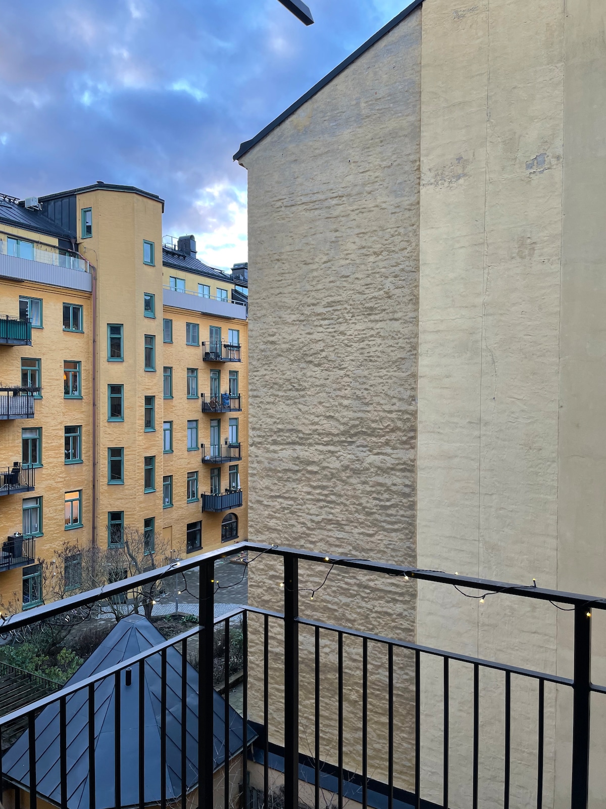 A view from the balcony captures neighboring buildings with yellow facades and balconies. The image showcases a textured wall of a close structure, with string lights adding a subtle glow, creating a cozy feeling in the shared courtyard space below.