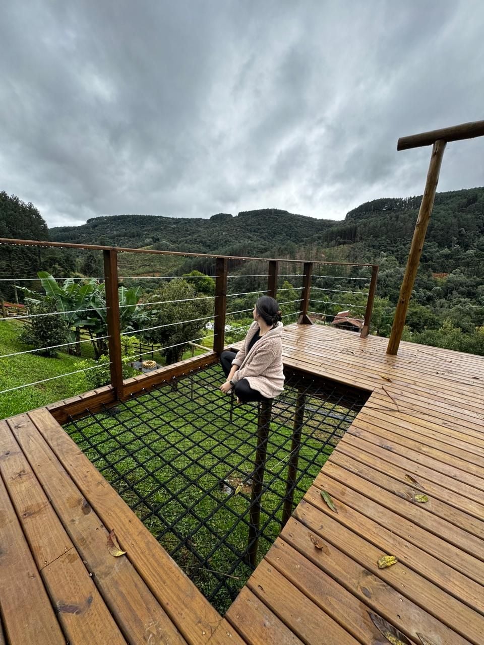 A wooden deck extends over the greenery, featuring a sturdy netted area where a person sits, facing the scenic hills. The sky is overcast, contributing to a serene outdoor environment surrounded by nature.
