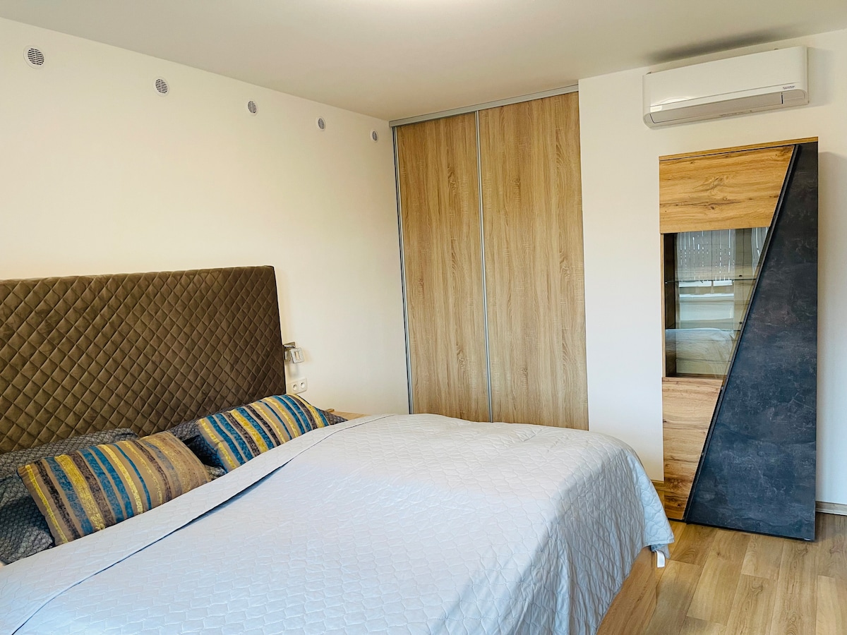 A modern bedroom displays a spacious bed with a textured headboard and patterned pillows. A wooden closet features sliding doors, while a stylish dresser with a glass section stands adjacent. Natural light filters in through the area, enhancing the room's calm atmosphere.