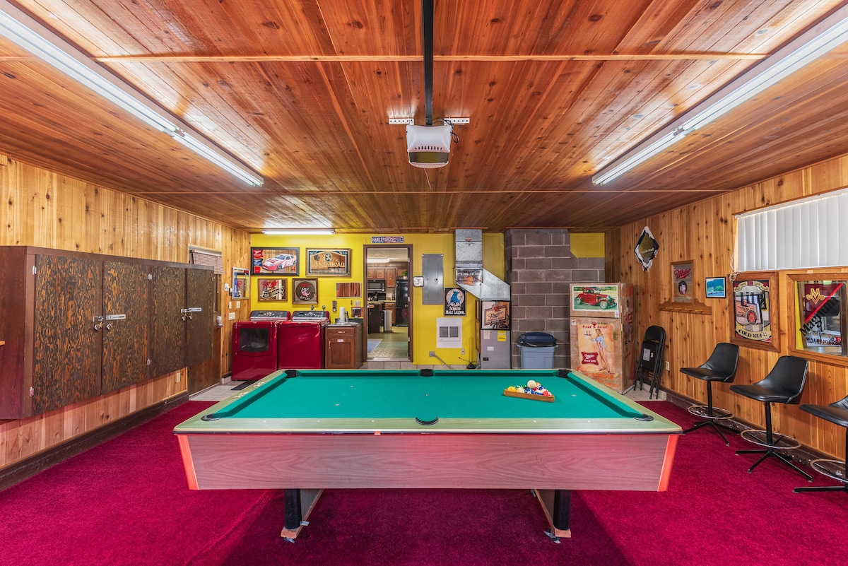 A spacious game room is highlighted by a green pool table, positioned centrally on deep red carpet. Wooden walls create a warm ambiance, while various framed artwork and memorabilia decorate the space. Bar stools are placed along a side, enhancing the recreational atmosphere.