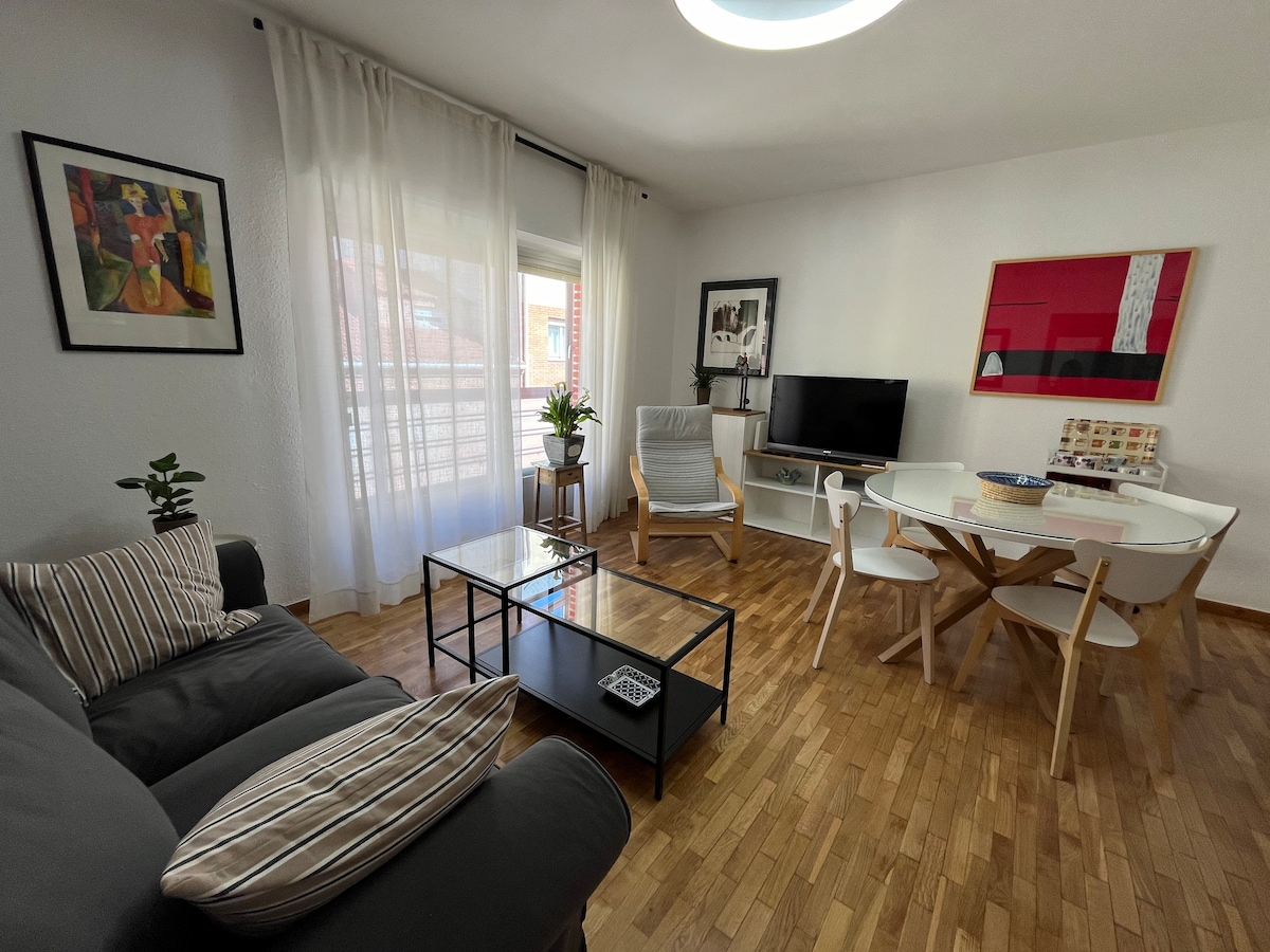 A well-lit living area is furnished with a gray sofa, a glass coffee table, and a round dining table surrounded by white chairs. A television sits on a wooden stand, while a potted plant adds a touch of greenery. Large windows draped with sheer curtains allow natural light to fill the space.