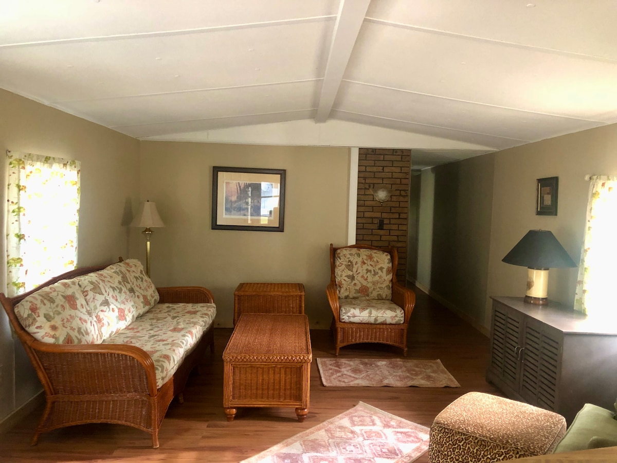 A welcoming living area features a pair of wicker seating arrangements, one with floral cushions and a matching coffee table. Soft light filters through sheer curtains on the windows, creating a warm ambiance in this cozy space. A lamp stands beside the sofa, adding to the comfort.