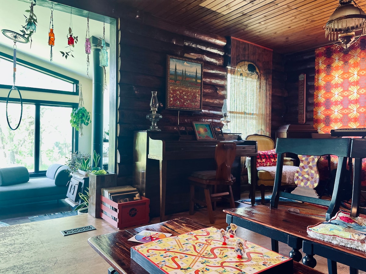 An inviting living area features rustic log walls and wooden flooring. A comfortable couch is positioned near a large window, while a vintage piano and colorful artwork enhance the space. A board game is set on a low table, inviting friendly gatherings.