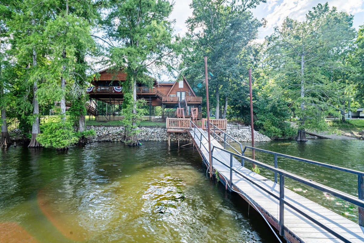 A wooden dock extends over the calm waters of the lake, surrounded by lush green trees. The two-story cabin is visible in the background, with a porch adorned with decorative accents. Sunlight filters through the trees, casting gentle reflections on the water's surface.