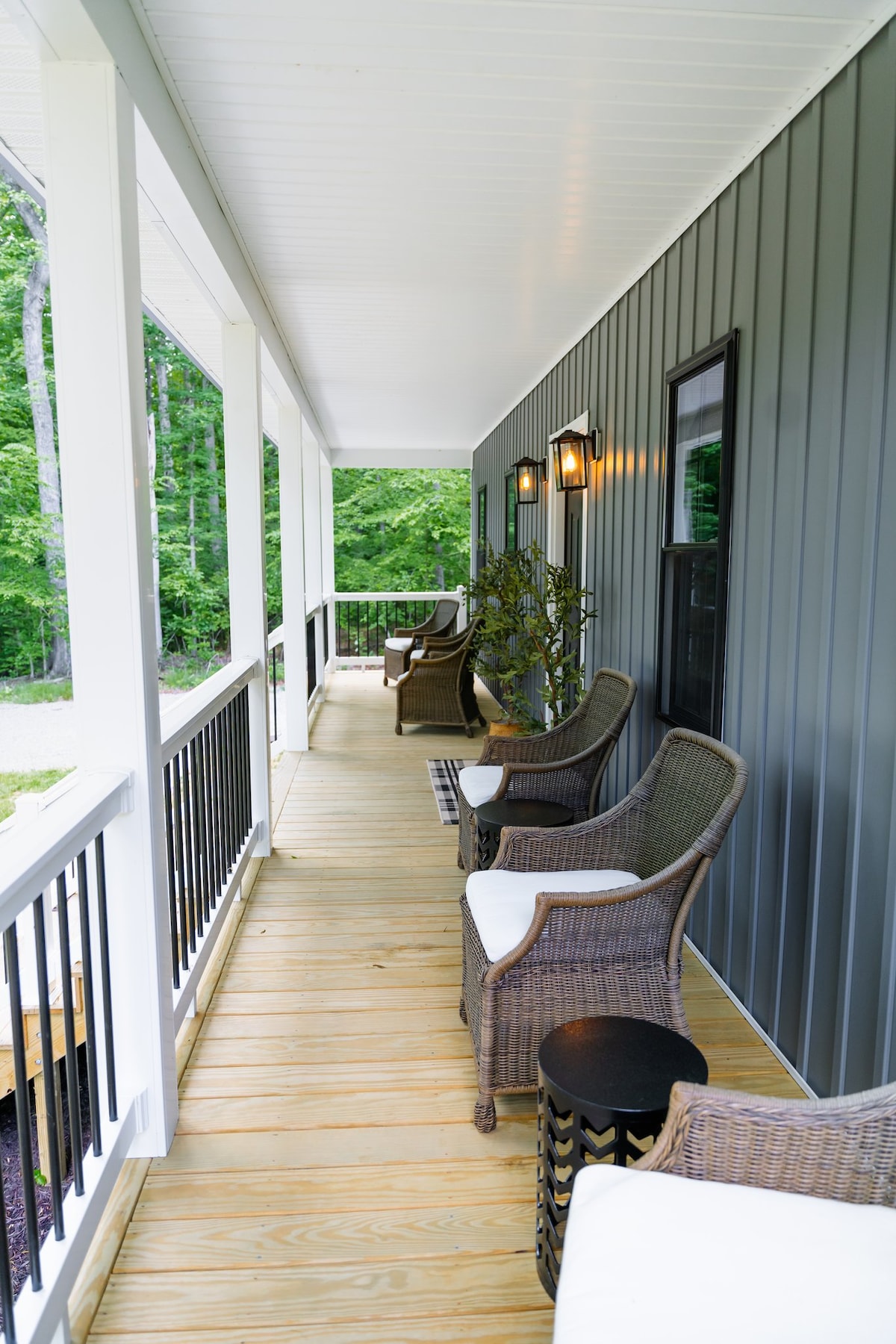A covered porch features several comfortable chairs set against a backdrop of lush greenery. Soft cushions are placed on the chairs, and side tables are positioned nearby. The area is well-lit with wall-mounted fixtures, offering a welcoming atmosphere for relaxation.