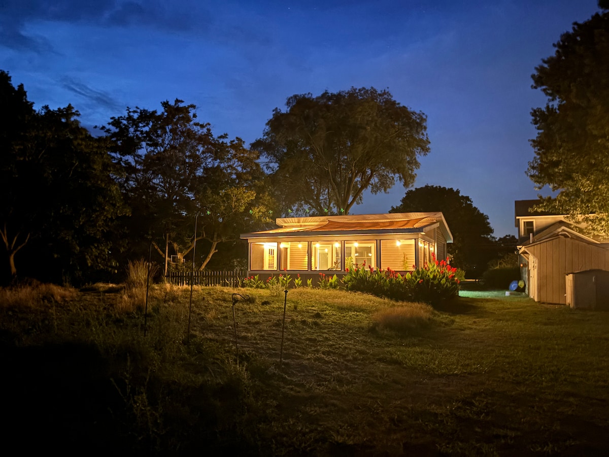 A tiny home is illuminated at night, surrounded by lush greenery and mature trees. The screened-in porch is visible, showcasing large windows that reflect warm light. The tranquil atmosphere is enhanced by the natural setting under a soft blue evening sky.