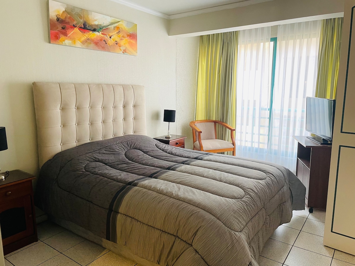 A spacious sleeping area features a soft, upholstered bed with a textured gray coverlet. Light filters through sheer curtains, illuminating the room. A stylish armchair and bedside lamps complement the decor, while an abstract artwork hangs above the headboard.