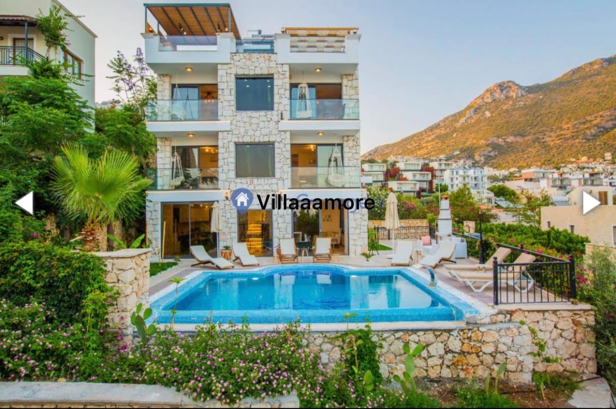 The exterior view of a multi-level villa is captured, showcasing a private pool surrounded by lush greenery. The stone façade complements large glass doors that invite natural light. Sunbeds and parasols are arranged beside the pool, with mountain scenery in the background.