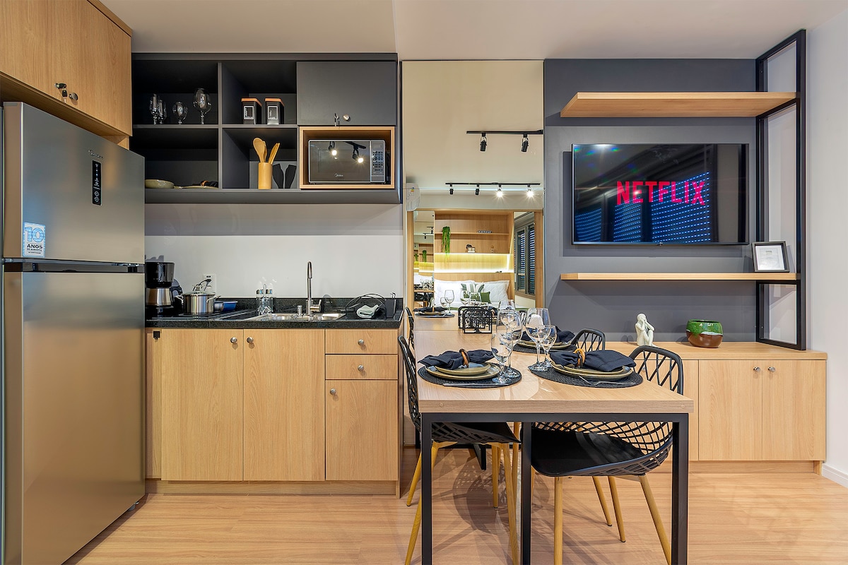 A modern kitchen and dining area is shown, featuring sleek wooden cabinetry and a well-equipped countertop. A dining table set for four is visible, complemented by stylish chairs. A large smart TV is mounted on the wall, with a cozy ambiance highlighted by soft lighting.