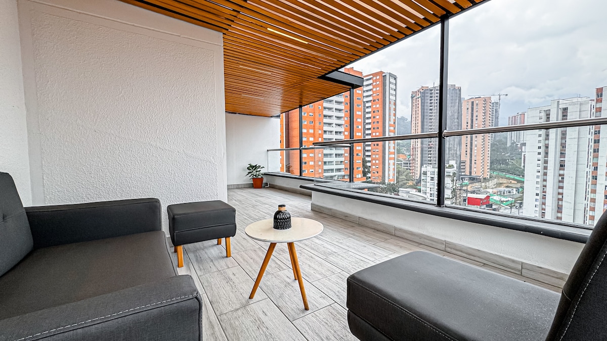 A covered terrace is presented with modern outdoor furniture, including a low table and two chairs. A nearby potted plant adds a touch of greenery. The expansive glass railing offers panoramic views of the cityscape, with various high-rise buildings visible in the background.
