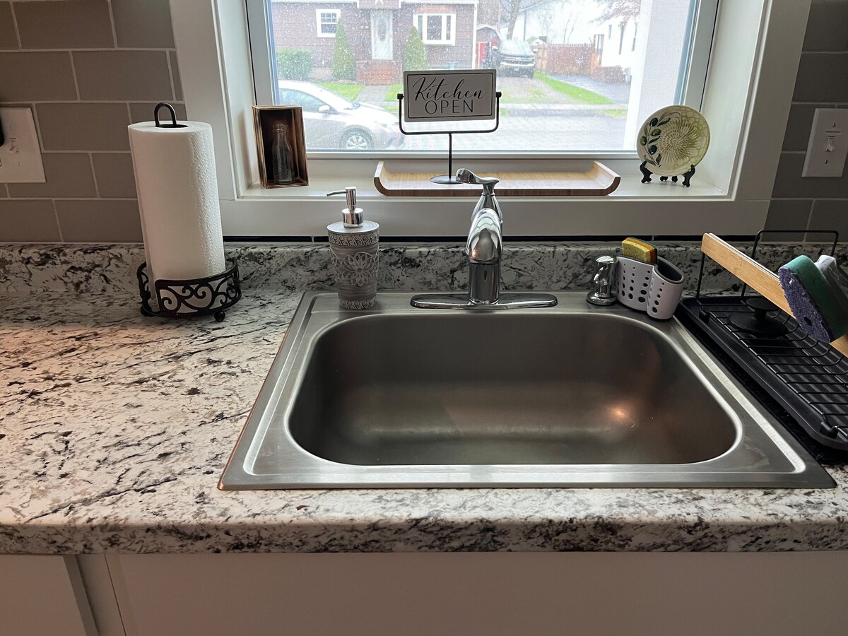 A spacious kitchen sink is set within a granite countertop. Natural light filters in from a window above, highlighting a clean design. Nearby, items like a dish rack, soap dispenser, and a paper towel holder are arranged neatly, contributing to a functional cooking space.