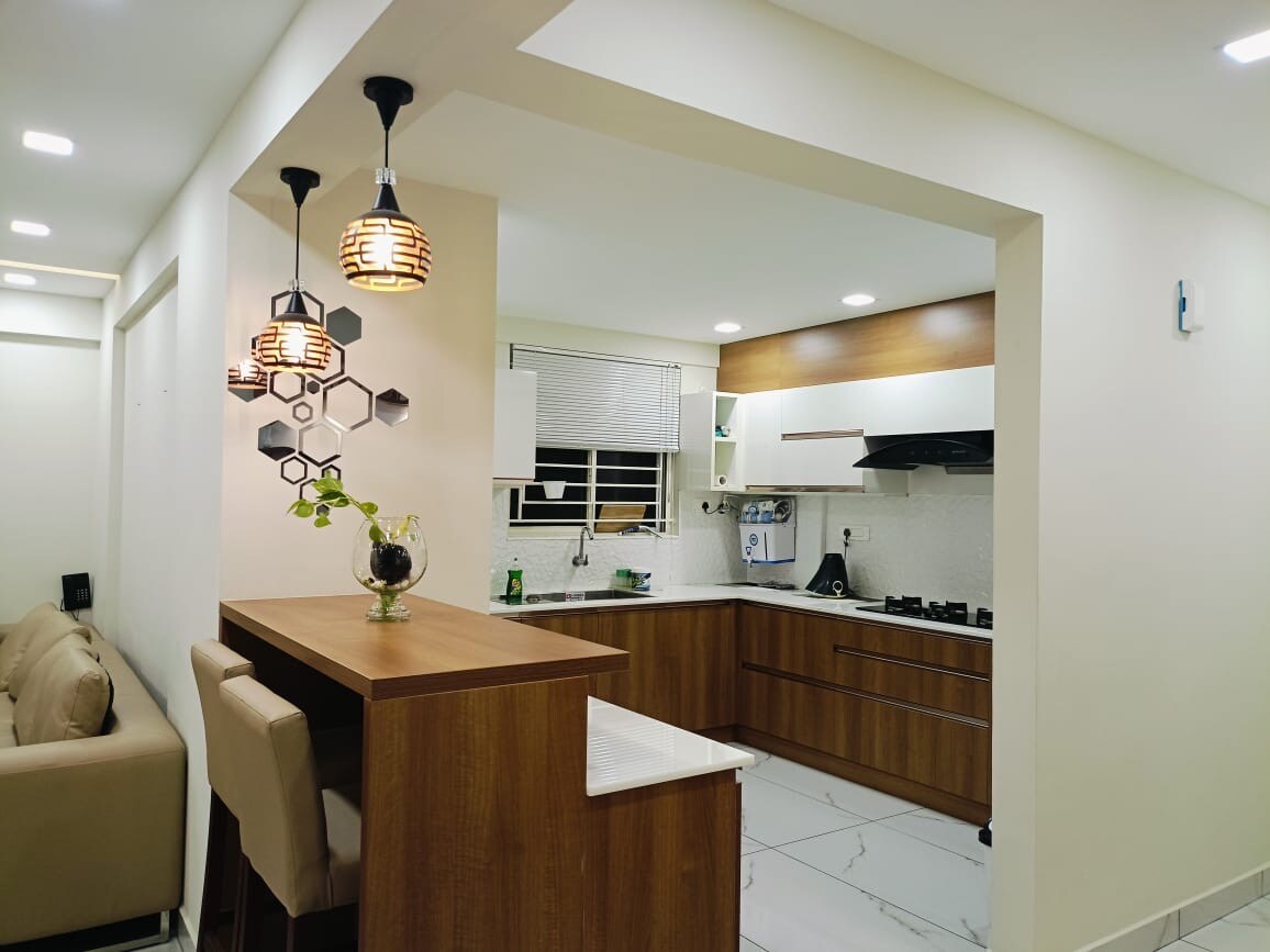 An open kitchen is displayed with modern cabinetry and sleek surfaces. Pendant lighting adds a touch of style to the space. A small bar area with seating complements the kitchen. Natural light filters through a window, illuminating the contemporary design.