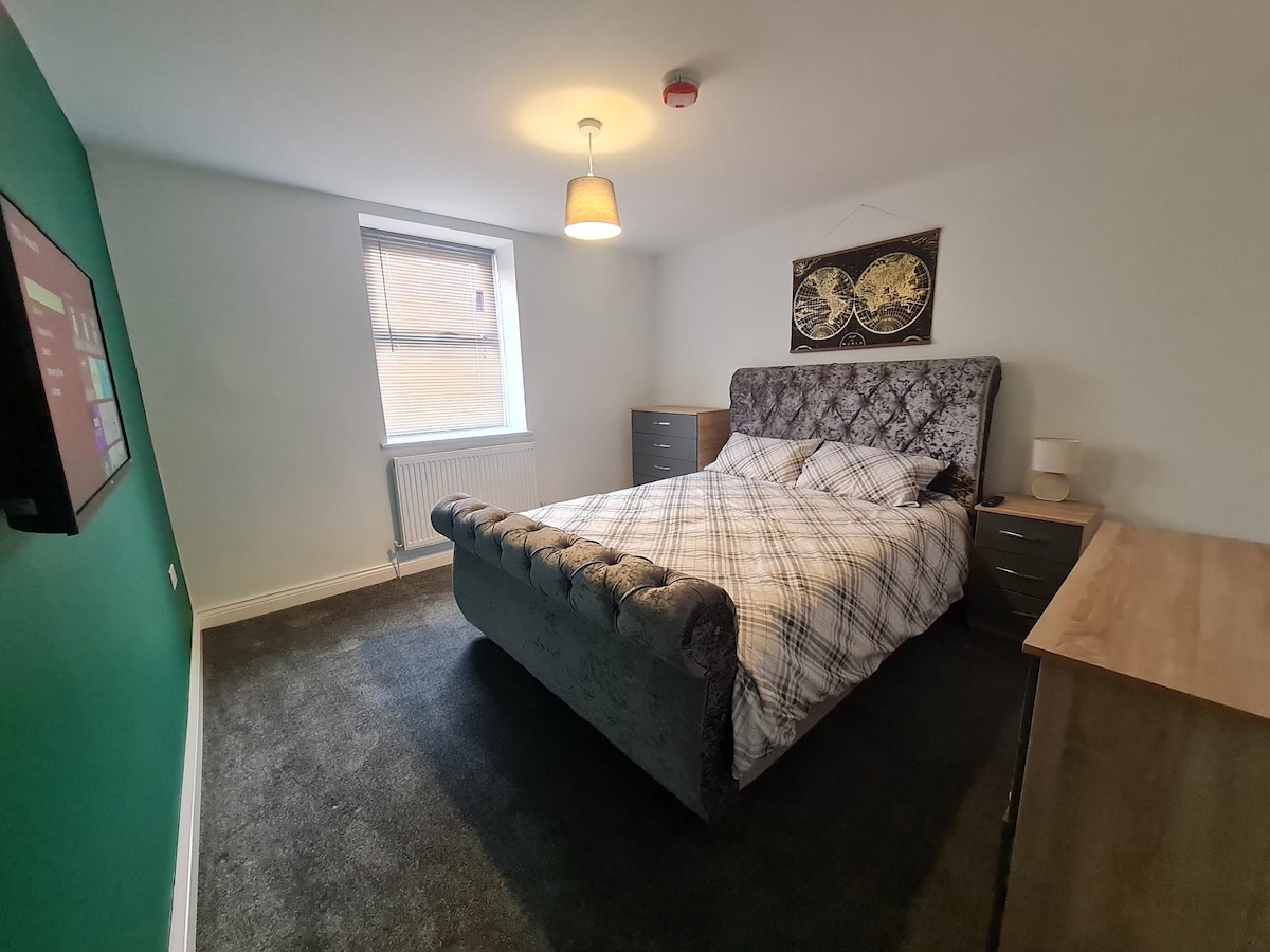 A comfortable bedroom features a plush dark gray bed with a stylish headboard, covered in a patterned blanket. A smart TV is mounted on the wall across from the spacious bed. Natural light filters through a single window, illuminating the room's neutral walls.