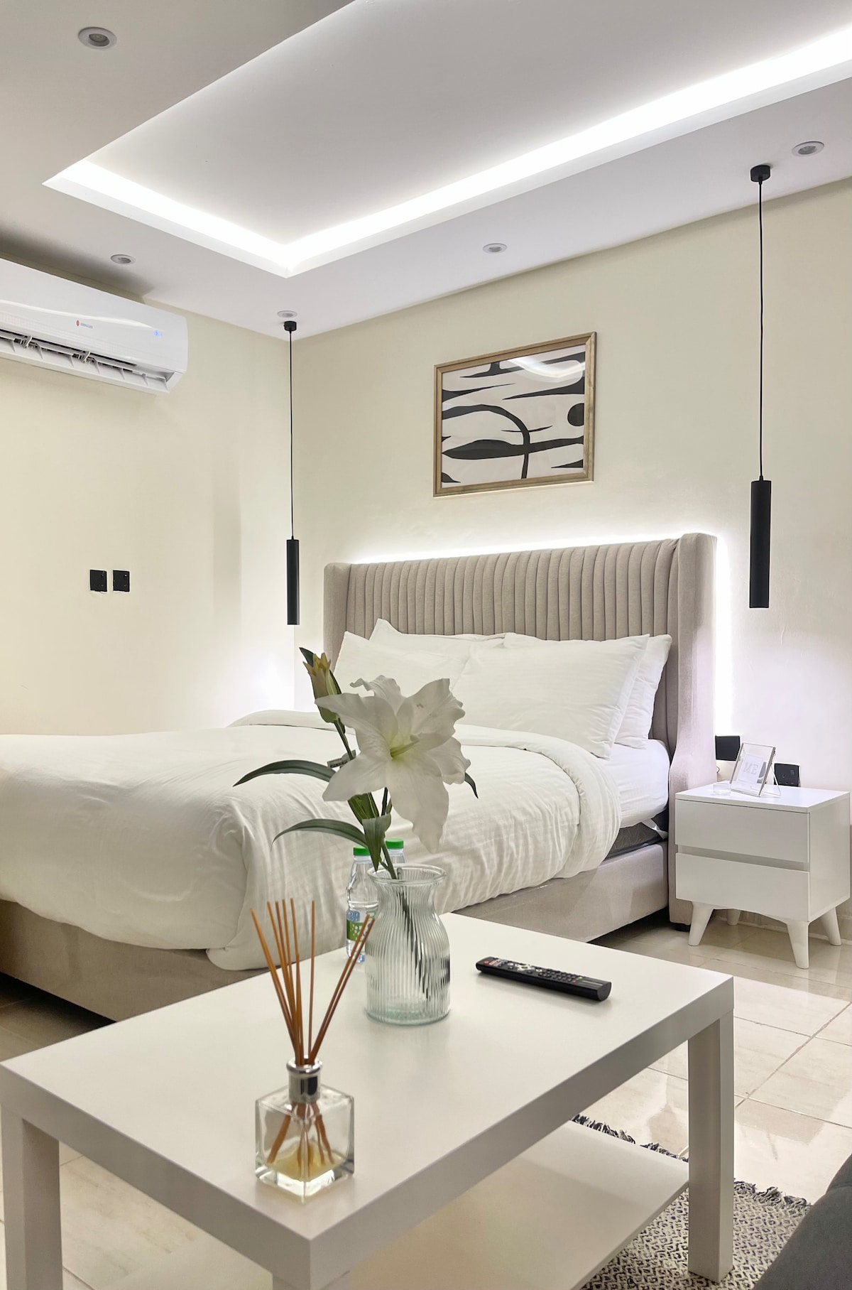 A serene sleeping area features a spacious bed with a soft headboard and muted bedding. A modern side table holds a remote control and a decorative vase with flowers. Pendant lights hang overhead, while artwork is displayed on the wall above the bed.