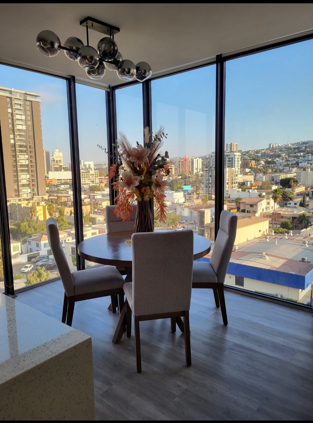 A dining area is featured, overlooking the cityscape through large glass windows. A round table is surrounded by four upholstered chairs, with a decorative floral arrangement as the centerpiece. Natural light floods the space, enhancing the modern design.