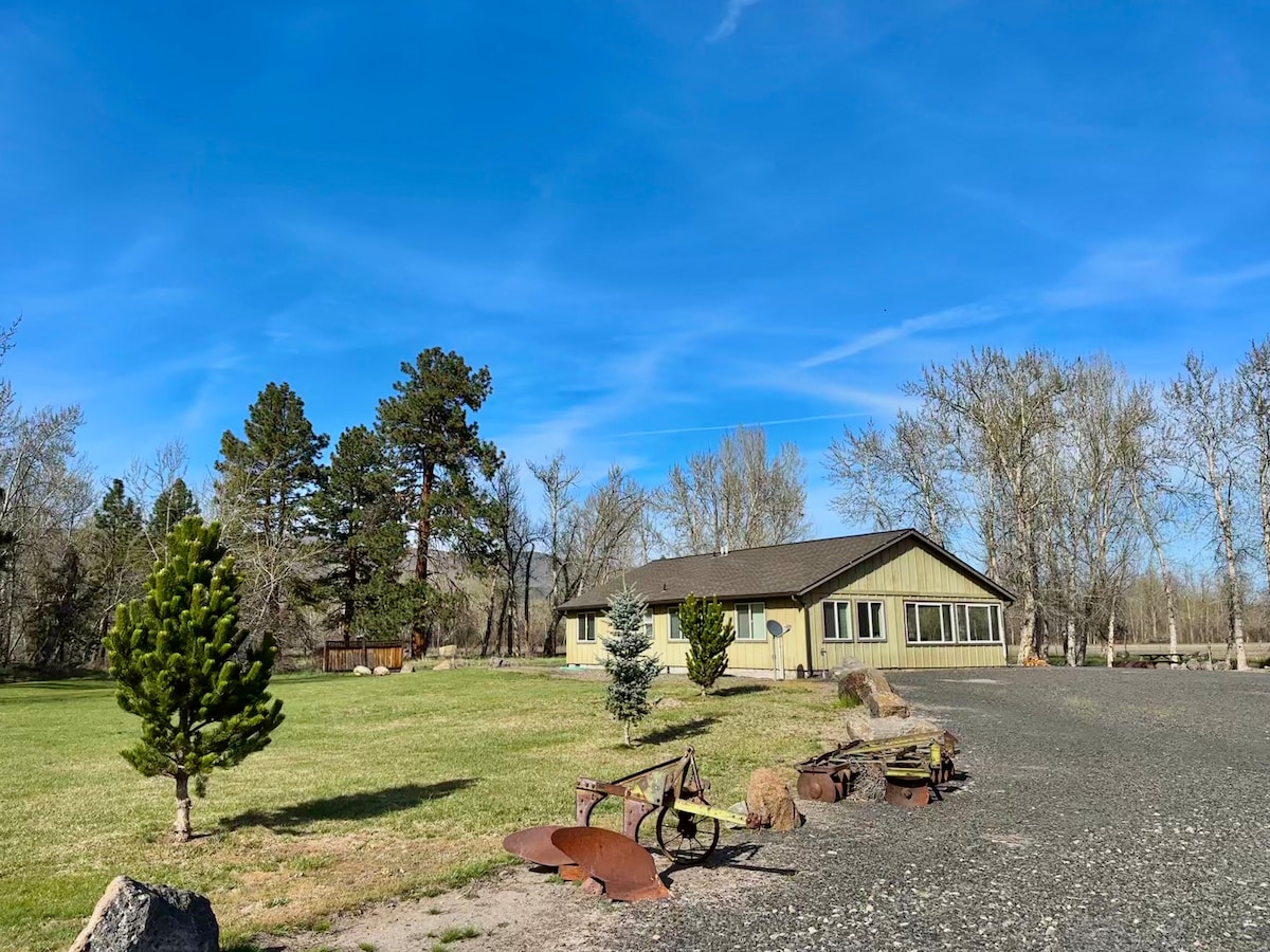 The ranch-style cottage is set amidst a grassy landscape, surrounded by trees. A gravel pathway leads to the entrance, with several large rocks and rustic tools placed decoratively nearby. The clear blue sky creates a serene backdrop for the peaceful environment.