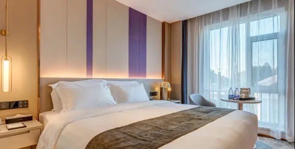 A modern bedroom features a spacious bed dressed in white linens, complemented by a decorative throw. Soft lighting is provided by sconces and bedside lamps. Large windows allow natural light to filter through sheer curtains, highlighting a small table with bottles of water and a chair.