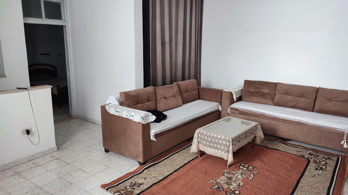 A spacious living area is furnished with two brown sofas arranged around a low table. An intricately patterned area rug covers the floor, while a curtain divides the room, adding a touch of privacy. Light enters through the nearby doorway.