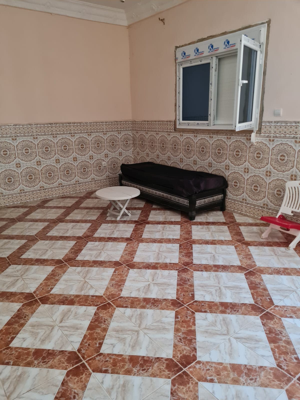 A spacious room features a patterned tiled floor in contrasting shades. A single bed with a dark covering is positioned against the wall, complemented by a small white table nearby. Natural light enters through a window, enhancing the room's bright and airy feel.
