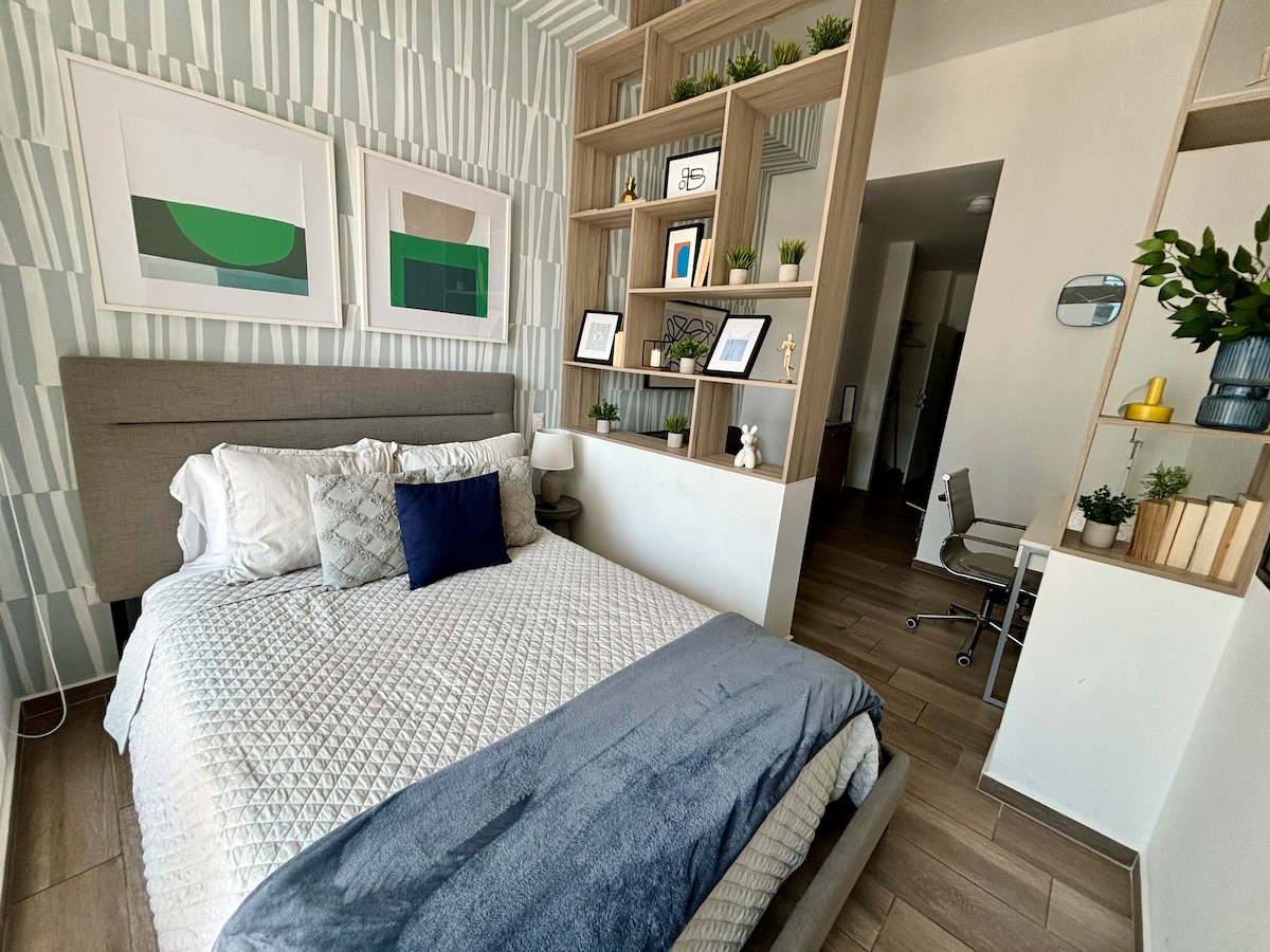A modern bedroom features a spacious bed with textured bedding and decorative pillows. Green and neutral wall art complements the stylish wallpaper. A minimalist desk and chair are positioned nearby, while shelves adorned with plants add a touch of greenery.
