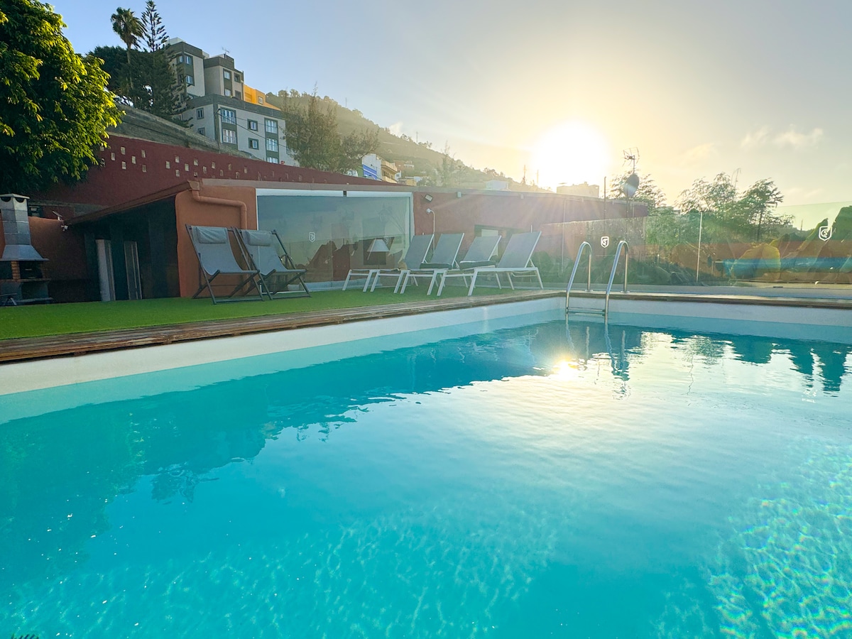 An outdoor swimming pool with clear blue water reflects the sunlight. Several lounge chairs are positioned nearby on a grassy area. A modern building can be seen in the background, framed by lush greenery and gentle hills.