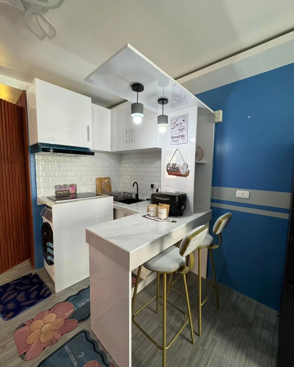 The kitchen area is equipped with modern white cabinetry and a marble countertop. A stovetop and a small coffee maker are visible, along with three bar stools positioned at the counter. A blue accent wall contrasts with the lighter elements, creating a contemporary look.