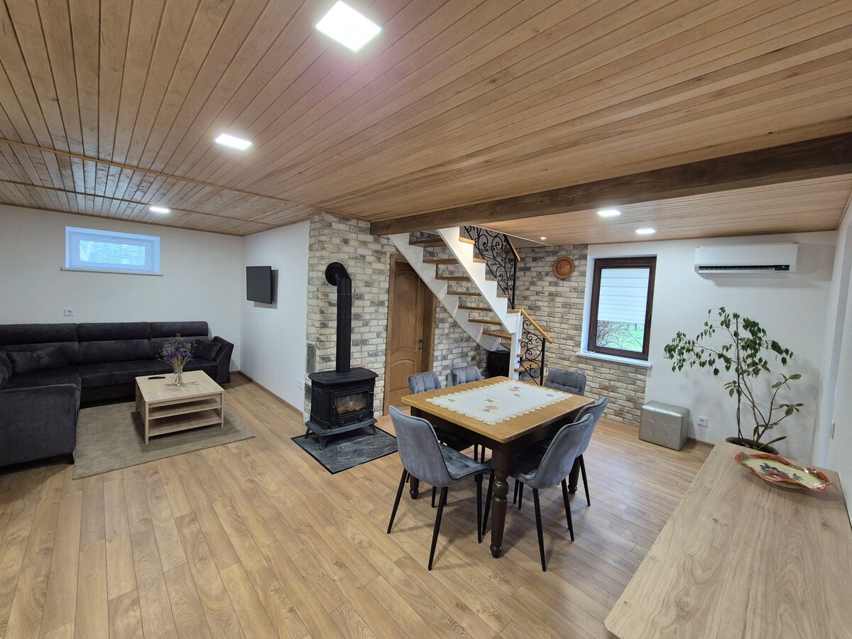 A spacious living area features wooden ceiling planks and light hardwood flooring. A comfortable sectional sofa is positioned nearby a rustic coffee table. A dining table with six chairs is centrally located, while a staircase leads to the upper level. A modern fireplace adds warmth to the space.