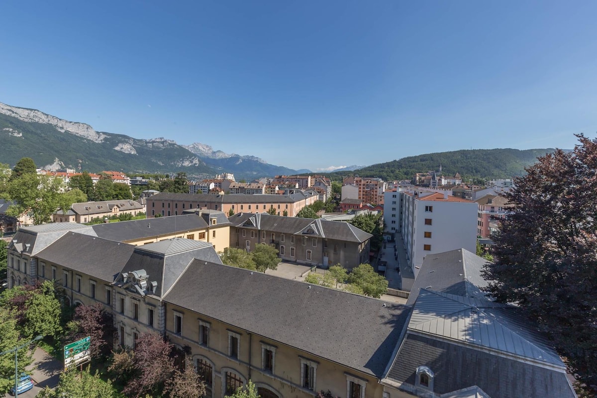 A panoramic view from an elevated perspective captures the surrounding architecture and green spaces of Annecy. The landscape features a mix of modern buildings alongside historical structures, with distant mountains creating a scenic backdrop against a clear blue sky.