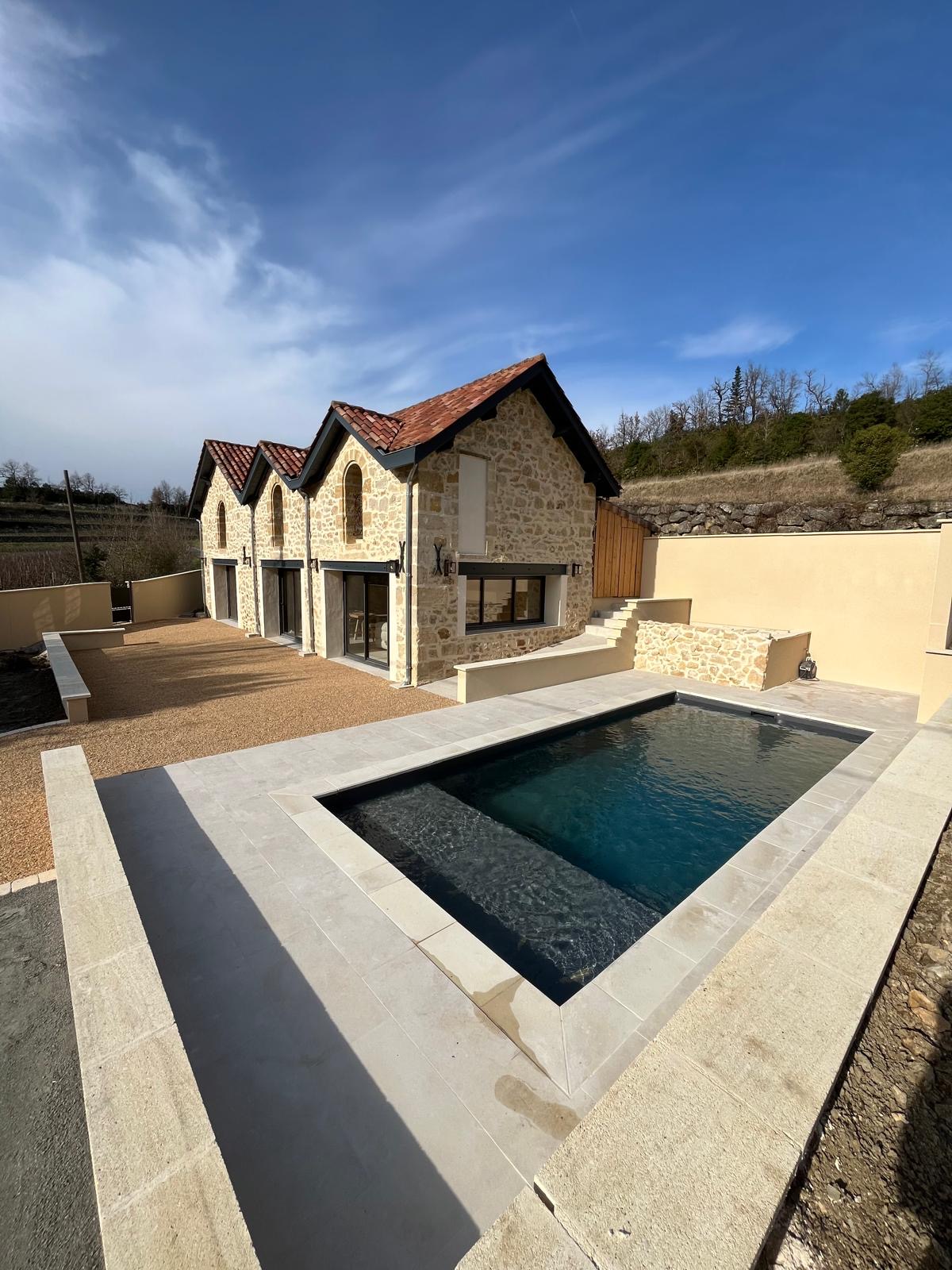 A villa with a natural stone facade is depicted, featuring a warm-colored roof and large windows. A calm, rectangular swimming pool sits in the foreground, surrounded by a patio area with a gravel finish, with gentle hills visible in the background.