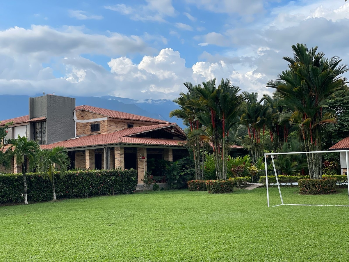 A spacious, two-story luxury house is surrounded by lush greenery, with palm trees and neatly trimmed hedges. A football goalpost is visible on the well-kept lawn, leading guests toward the inviting entrance of the property against a backdrop of distant mountains.