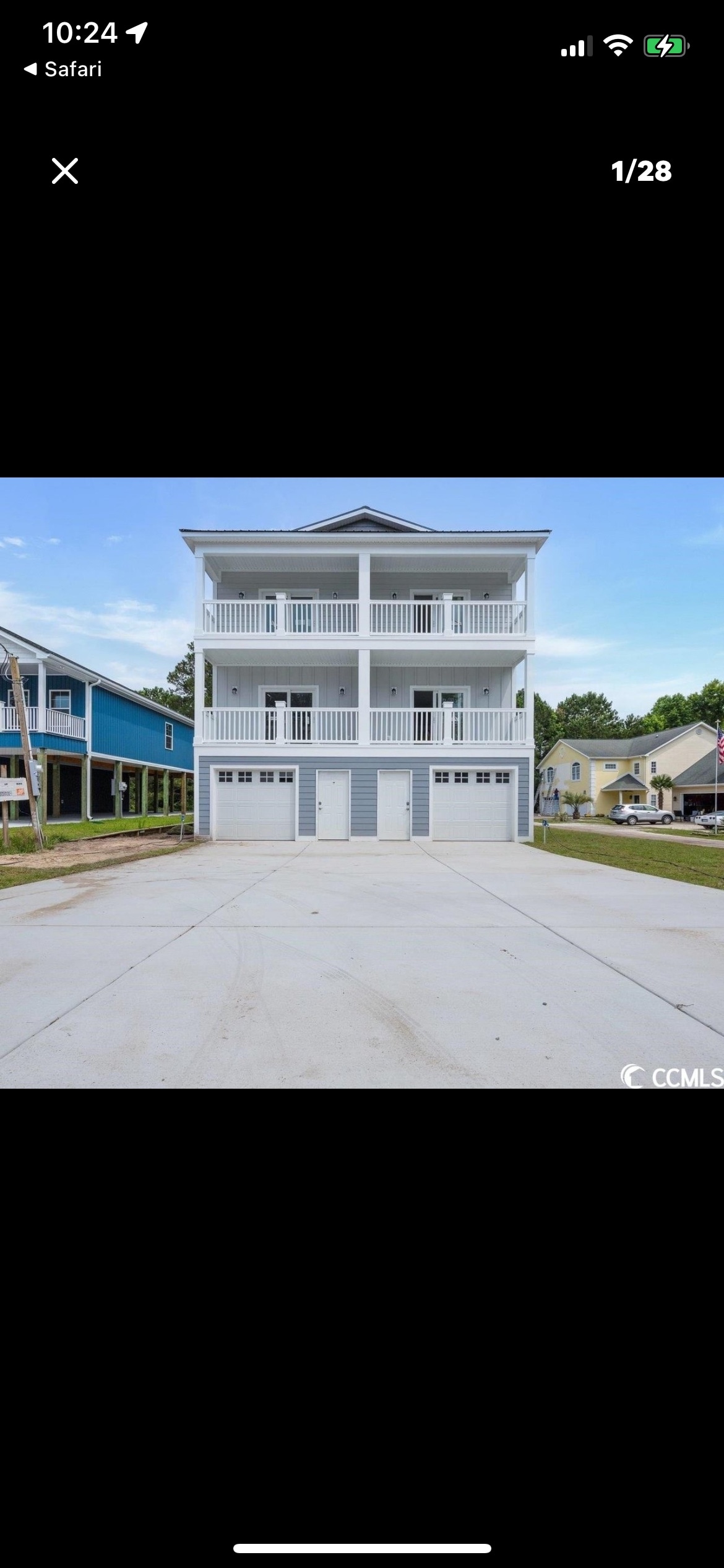 A three-story duplex is displayed, featuring a modern architectural design with two balconies and a wide driveway below. The exterior is painted in a soft gray, complemented by white railings. Two garage doors are visible on the ground level, enhancing the structure's functionality.
