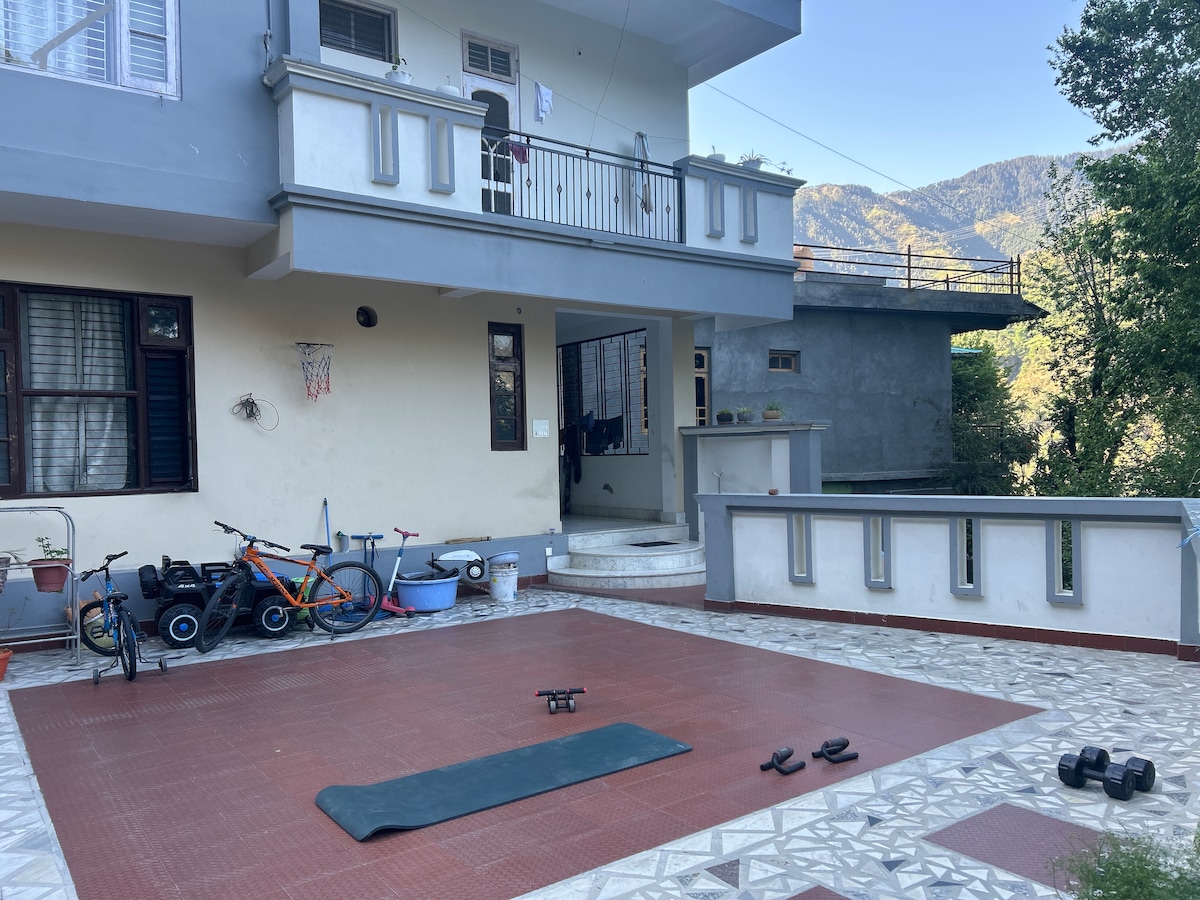 An outdoor space features a large patio with exercise equipment, including a yoga mat and weights. Nearby, a basketball hoop is mounted on the wall. Bicycles are parked to one side, with greenery and mountains visible in the background.
