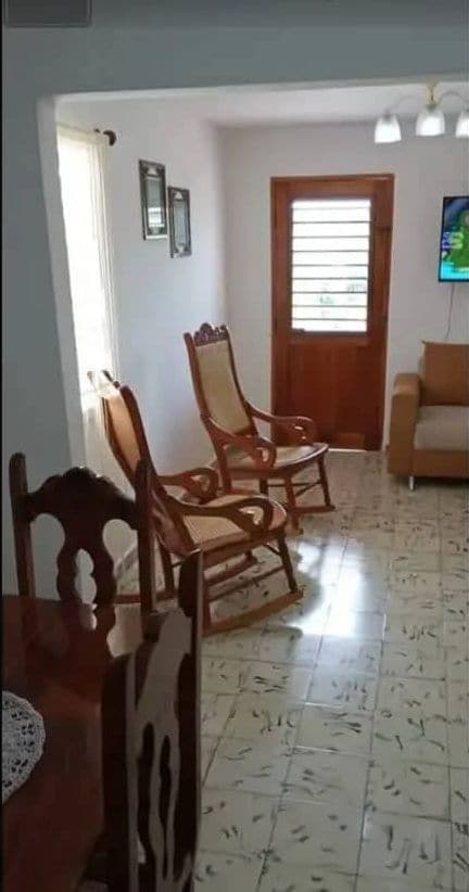 A living area featuring two rocking chairs and a small sofa. Light-colored walls complement the space, while natural light enters through a window beside the entrance door. Decorative frames are mounted on the walls, adding character to the room.