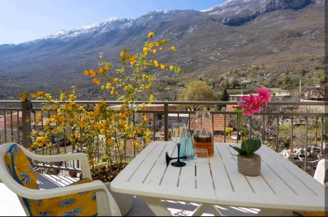 A balcony is showcased with a table and two chairs. A potted pink flower adds a touch of color, accompanied by a bottle and glasses. Vibrant yellow flowers bloom nearby, while the background features a mountainous landscape adorned with snow-capped peaks.