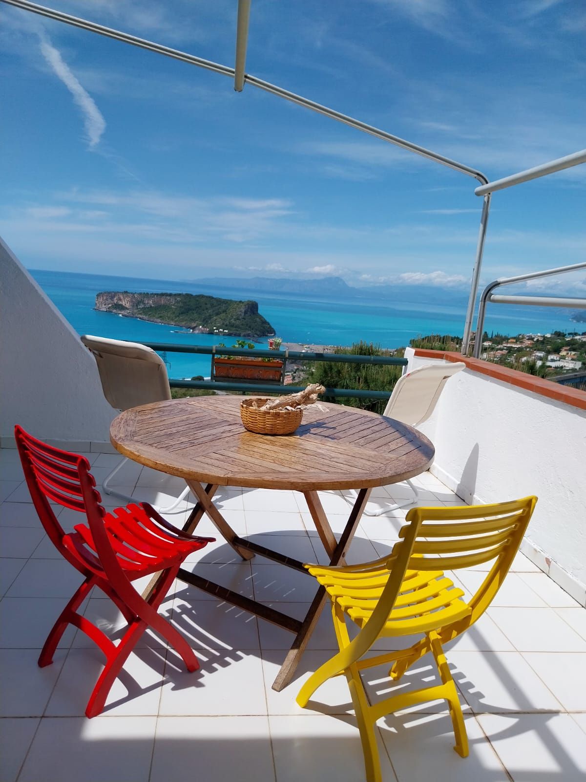 A quaint outdoor terrace is equipped with a round wooden table, surrounded by yellow and red folding chairs. A basket sits at the center of the table. The view showcases a panoramic scene of blue waters and distant landforms under a clear sky.