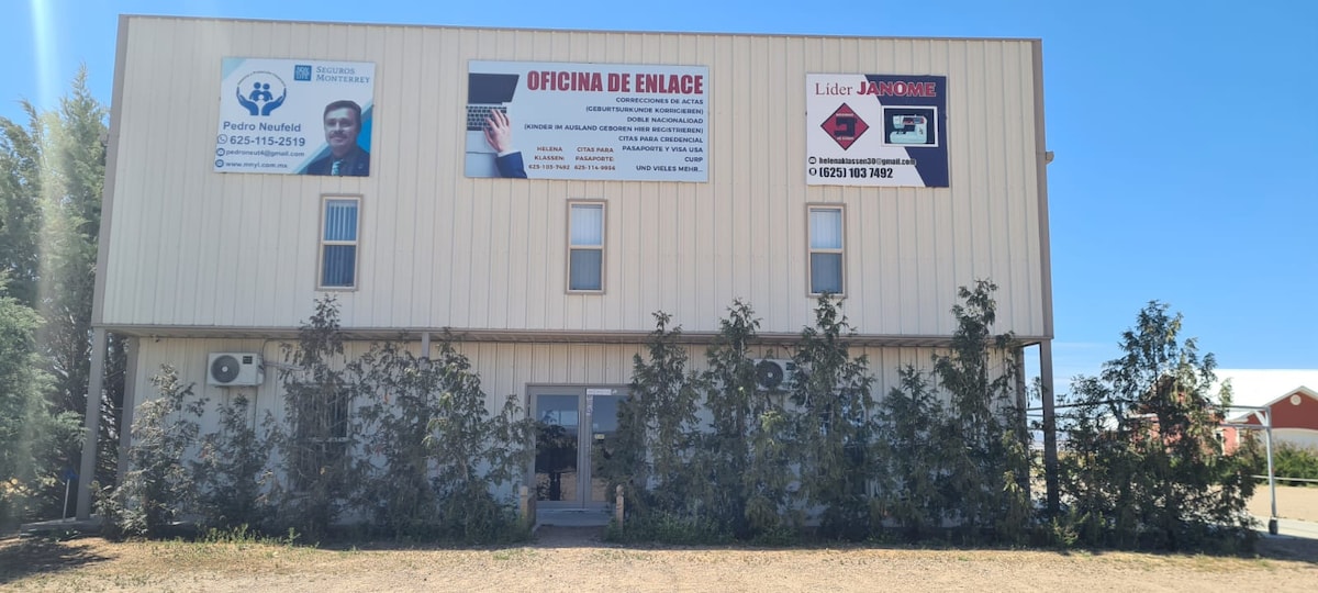A light-colored two-story building is displayed, featuring a simple façade with signs advertising various services. Small shrubs are seen along the front, complemented by a gravel surface leading to the entrance. Windows are positioned on both levels, allowing for natural light.
