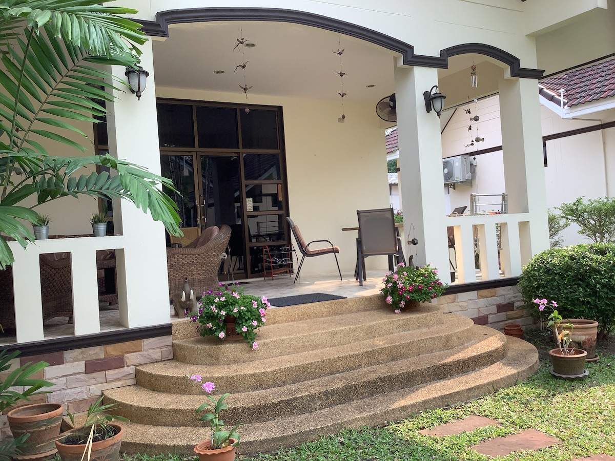 A covered entryway is framed by a spacious porch featuring multiple seating areas. Decorative plants in pots are arranged along the steps, leading up from a well-maintained lawn. Light filters softly through large windows, contributing to an open and inviting feel.