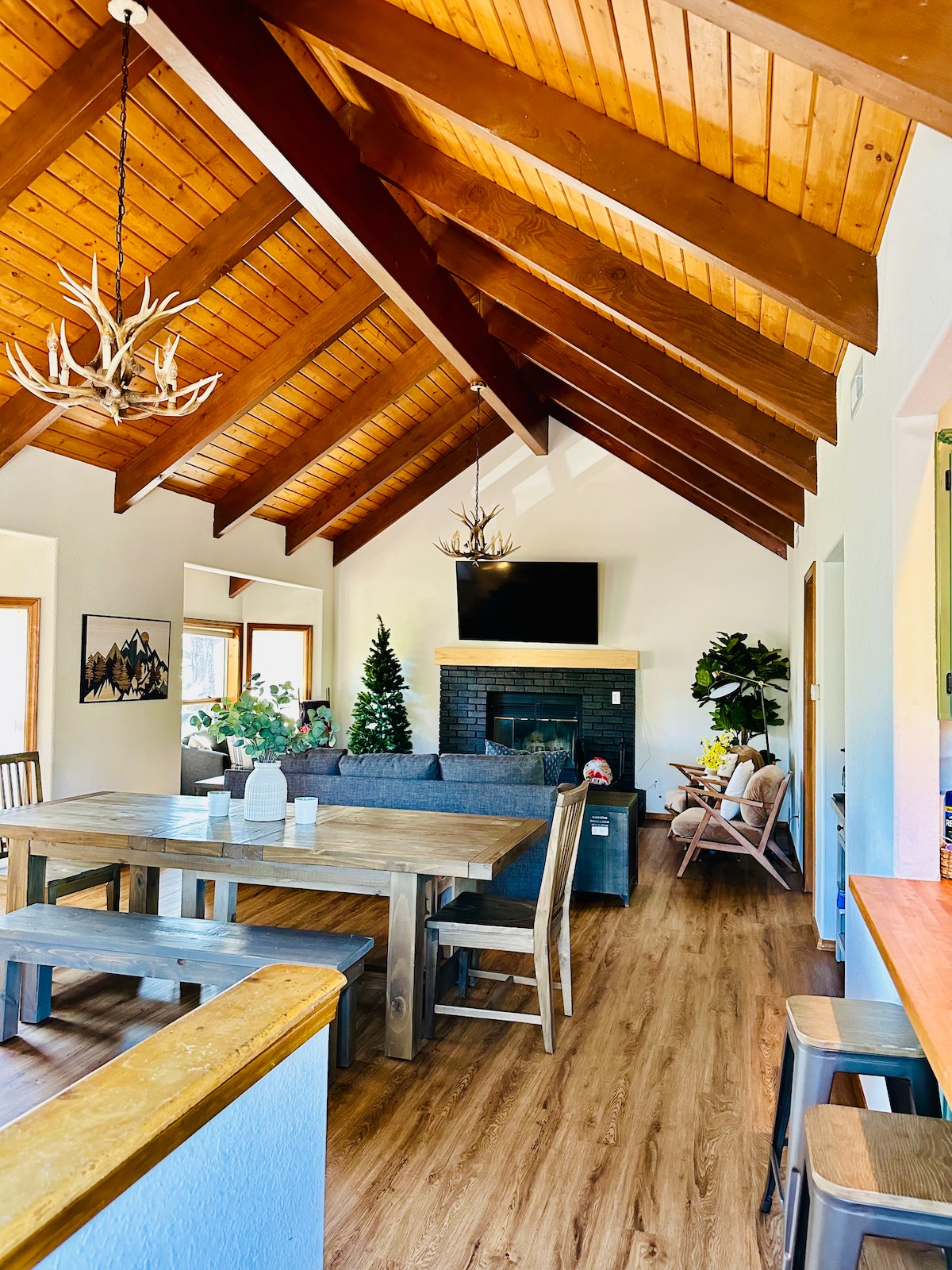 An inviting living area features a high wood-beamed ceiling with a light fixture, accompanied by a large window and a stone fireplace. A wooden dining table is positioned nearby, with seating for several. A cozy seating arrangement includes a sofa and accent chairs, complemented by indoor plants.
