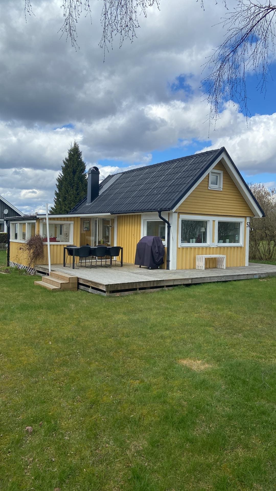 A charming yellow cottage with a black roof is situated in a grassy area. A spacious wooden deck features seating arrangements and is complemented by a tree in the background. Soft clouds are seen in the sky above, contributing to a serene outdoor setting.
