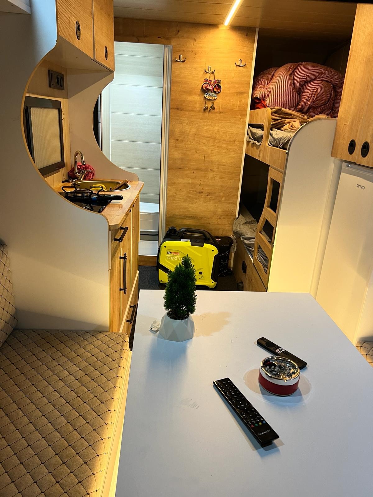 An interior view of the caravan displays a practical layout, featuring a compact table accompanied by seating. The kitchenette is visible, with essential appliances and storage. In the background, a bunk area is seen, along with a bathroom space, all enhanced by warm wooden finishes.