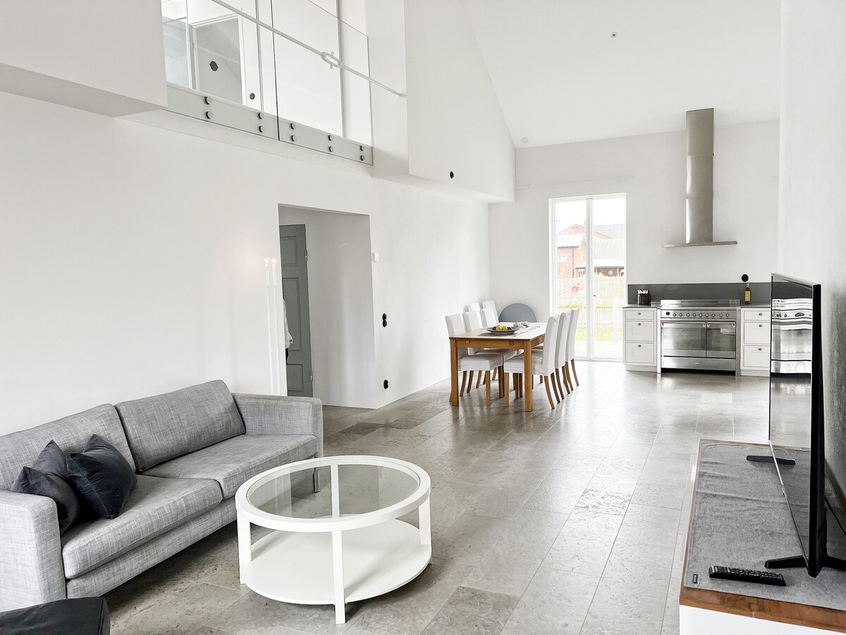 An open-plan living area features a gray sofa and a circular coffee table, with light-colored tile flooring. A dining table surrounded by white chairs is positioned beside a fully equipped kitchen. Large windows allow natural light to fill the space, enhancing its airy atmosphere.