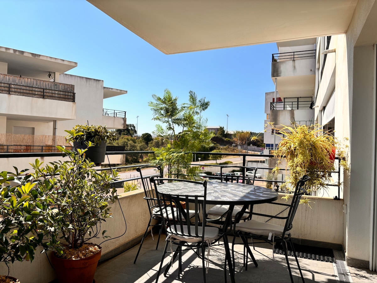 A balcony features a round table surrounded by four black chairs. Potted plants are located on the table and around the railing, while sunlight brightens the area, creating an open and airy feel. Nearby buildings and greenery are visible in the background.