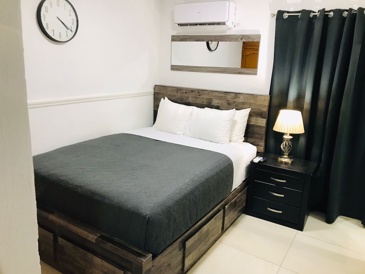 A spacious bed is framed by a wooden headboard, featuring crisp white linens and a dark grey blanket. A bedside table with a lamp is positioned next to the bed, while dark curtains cover the window, allowing for privacy.