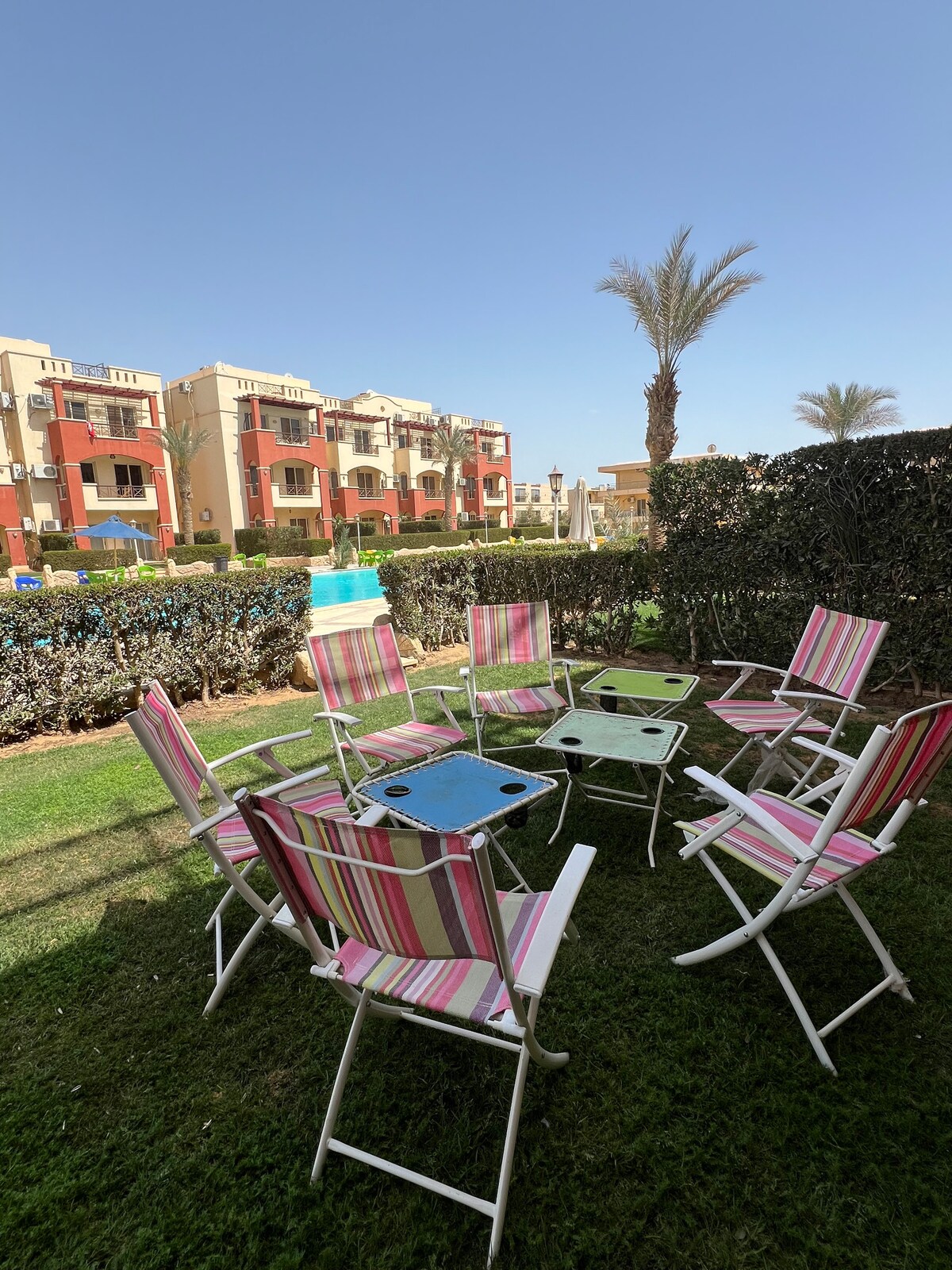 A group of colorful striped chairs surrounds a small table on a well-maintained lawn. Nearby, palm trees and decorative shrubs frame the scene, while low-rise buildings are visible in the background under a clear blue sky.